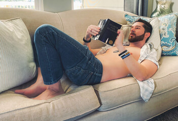 attractive, sexy man or male model with beard laying down inside home reading book shirtless and...