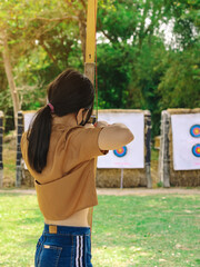 Back view of Asian girl wear face mask aims archery bow and arrow to colorful target in shooting range during training and competition. Exercise and concentration with outdoor archery.Archery for teen © JinnaritT