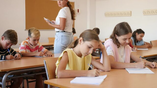 Young girl sitting at desk with her classmates during lesson in school and writing in copybook. 
