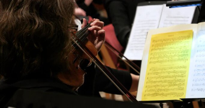 Close up of a female violin player performing with a wooden violin with orchestra during a concert with classical music to mozart requiem with view of other violin players and sheet music. Slow Motion