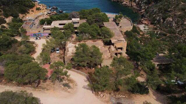 Abandoned hotel on cliff top at Cala d&rsquo;en Serra Beach, Spain with graffiti, trees, roads and ocean cove in background - Drone aerial flyover footage.