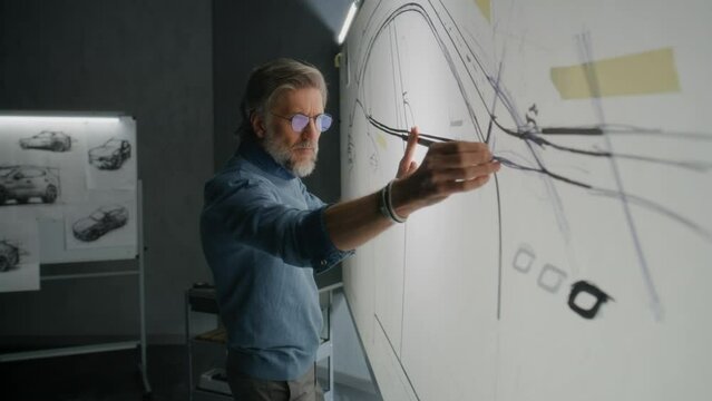 Senior automotive male designer creating sketch with duct tape on paper whiteboard, developing futuristic electric car future design. He working in modern car design development studio.