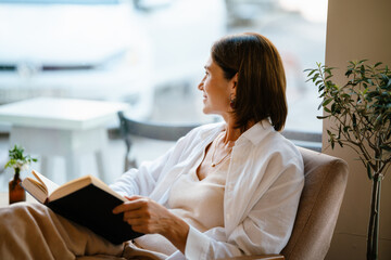 White mature woman smiling and reading book while sitting in cafe