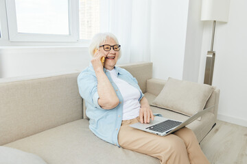 a joyful elderly lady is talking on a smartphone sitting on a sofa in a bright apartment holding a laptop on her lap smiling broadly