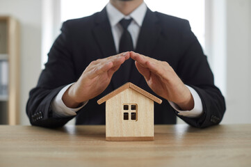 Insurance agent in a suit covering a small wooden toy house with his hands like a roof. Cropped shot, close up. Home, property, safety, protection, house insurance concept