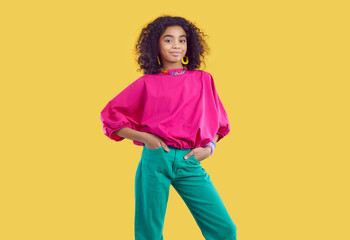 Studio portrait of beautiful stylish dark-skinned curly preteen girl isolated on yellow background. Ethnic girl in fashionable colored teenage clothes and with accessories smiling at camera.