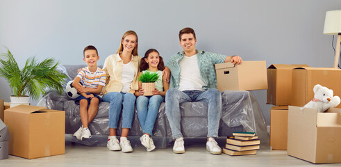 Fototapeta na wymiar Mortgage and buying property. Happy young family with children sitting on sofa between cardboard boxes in their new apartment. Concept of buying your own house and moving. Web banner.
