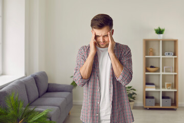 Stressed young man having a simple headache or a severe migraine. Tired, nervous, worried man standing in the living room, feeling pain in his head and touching his temples