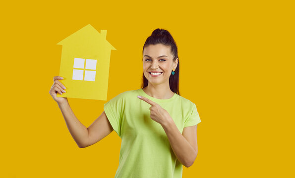 Happy beautiful young woman standing against bright yellow studio background, pointing at symbolic paper house that she is holding, looking at camera and smiling. Real estate, buying new home concept
