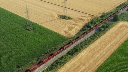 Aerial view, drone orbit, panning lateral over long composition, empty freight railroad train, passing cultivated plots. Mature wheat, corn, sunflower in green vegetation, divided agricultural fields