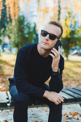 Fototapeta premium A lonely young man in a black sweater sits in the park on a bench and talks on the phone. A warm autumn day