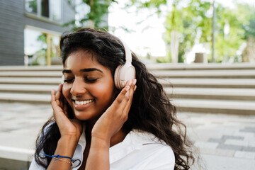 Young indian woman smiling and using headphones while sitting outdoors