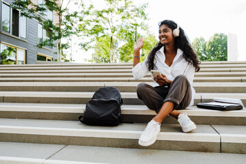 Young indian woman using cellphone and waving to someone outdoors