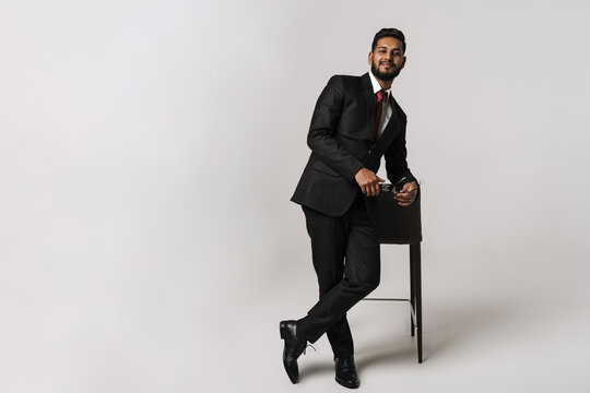 Young indian man wearing suit posing with glasses and chair