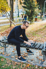 Fototapeta premium A young man sits in the park on a bench and works on a laptop. A warm autumn day. Remote work