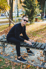 Fototapeta premium A young man sits in the park on a bench and works on a laptop. A warm autumn day. Remote work