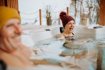 Senior couple in kintted cap enjoying together outdoor bathtub at their terrace during cold winter day.