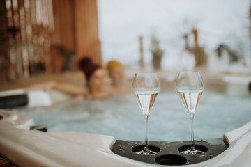 Senior couple in kintted cap enjoying together outdoor bathtub with glass of wine, at their terrace during cold winter day.