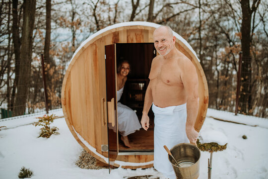 Senior woman in towel with her husband entering to outdoor sauna during cold winter day.