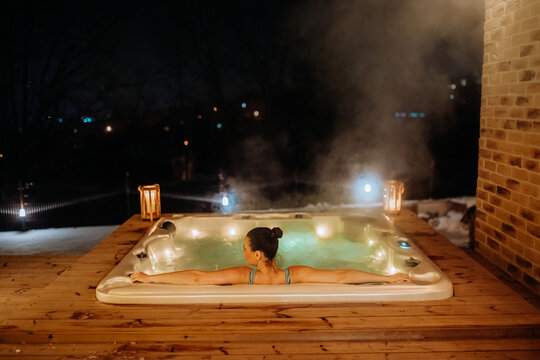 Rear view of young woman enjoying outdoor bathtub in her terrace during cold winter evening.