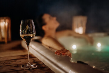 Young woman enjoying outdoor bathtub with glass of wine at her terrace during cold winter evening.