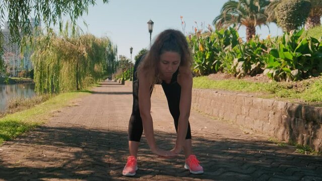 Young beautiful caucasian lady wearing sportwear bend her body and reach hand to touch ground floor. Stretching body, prepare for exercise outdoors. Fitness in park of resort town.