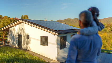 Rear view of dad holding her little girl in arms and looking at their house with installed solar...