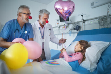Happy doctors with clown red noses celebrating birthday with little girl in hospital room.
