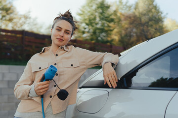 Young woman holding power supply cable from her electric car, prepared for charging it in home,...