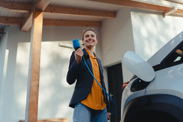 Young woman holding power supply cable from her electric car, prepared for charging it in home,...