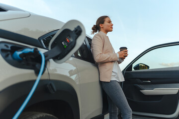 Young woman with cup of coffee waiting while her electric car charging, sustainable and economic...