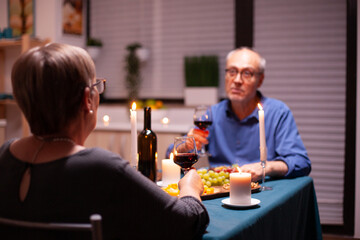 Woman having a romantic dinner with husband in kitchen in the evening holding glass of wine. Senior couple sitting at the table in dining room , talking, enjoying the meal, celebrating their