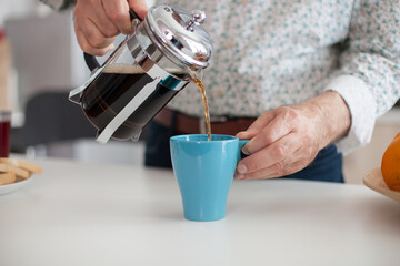 Senior man pouring hot coffee from french press during breakfat in kitchen. Elderly person in the...