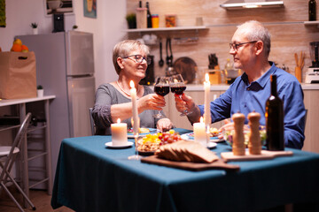 Fototapeta na wymiar Candles burning on kitchen table and old couple toasting with glasses for red wine for their relationship. Happy cheerful senior elderly couple dining together in the cozy kitchen, enjoying the meal.