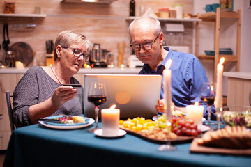 Pensioner woman using credit card to pay for romantic dinner using laptop in kitchen for her and husband. Old people sitting at the table, browsing, using the technology, internet.