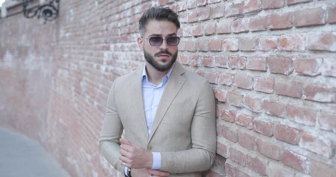 project video of elegant bearded man arranging suit sleeves, looking to side and laying on a bricks wall outside in a medieval city from Romania