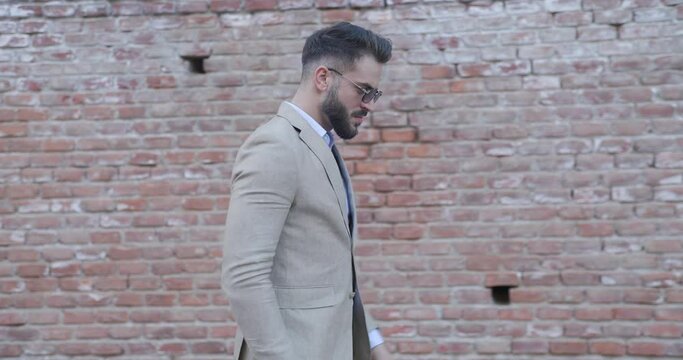 elegant stylish man arranging suit, looking around while walking in front of bricks wall and making a gun gesture with his hands outside in an old town from Romania