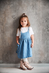 Full length of adorable little girl in denim dress standing against the wall in new home. Cute female child keeping hand in pocket and looking at camera.