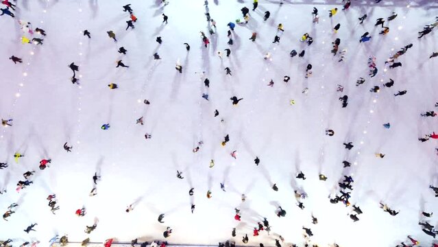 People skating on ice skating rink outdoors on winter night top view. Aerial drone. Luminous glowing garlands lamps lighting. Long shadows. New Year Christmas celebration holidays recreation Rising up