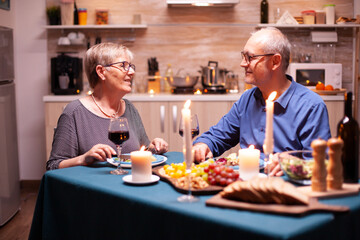 Fototapeta na wymiar Having a conversation during din romantic dinner. Happy cheerful senior elderly couple dining together in the cozy kitchen, enjoying the meal, celebrating their anniversary.