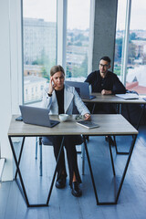 A female manager sits at a desk using a laptop in a modern office with a colleague in the background. Working atmosphere in an office with large windows.