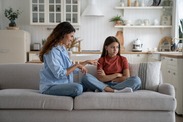 Shy little teenager girl turns away from smiling mother who is trying to communicate. Cheerful kind Caucasian woman sits on sofa in spacious apartment touches daughter wanting to cheer up child