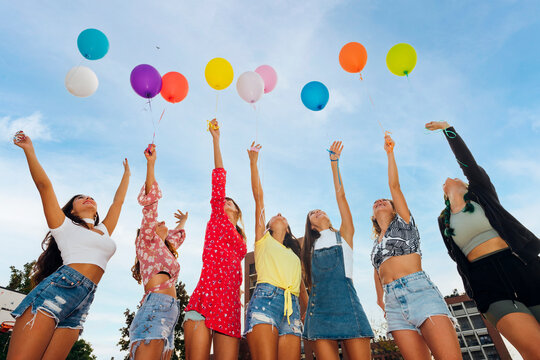 Friends with hands raised holding colorful balloons under blue sky