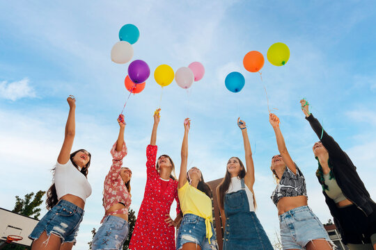 Happy friends with hands raised holding colorful balloons under blue sky