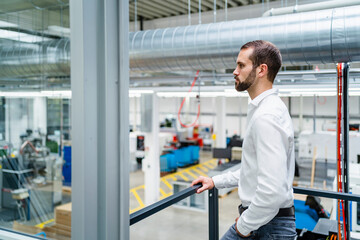 Serious young businessman with hand in pocket standing by railing in factory