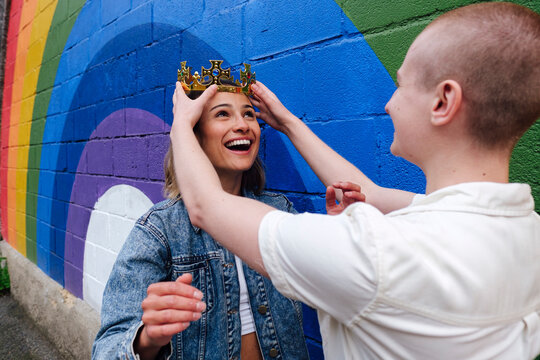 Non-binary person adjusting crown on lesbian woman's head by rainbow wall