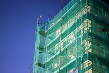 Multistory building facade covered with green protective construction mesh on blue sky background. Construction, reconstruction of residential buildings, house renovation, skyscrapers in big city.