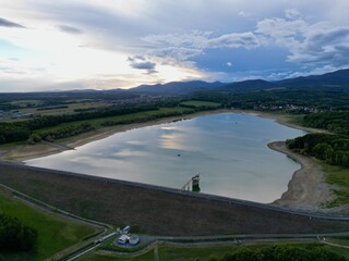 Fototapeta premium Flight over the Michelbach lake dam (Upper-Rhine, Alsace, France) by drought and cloudy weather in summer with a very low water level, in the evening at sunset with village and mountains in background