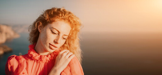 Close up shot of beautiful young caucasian woman with curly blond hair and freckles looking at camera and smiling. Cute woman portrait in a pink long dress posing on a volcanic rock high above the sea