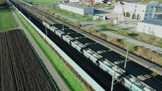 A freight train carrying gravel through an industrial area is followed by a drone flying next to the train tracks.